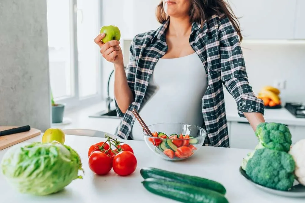 Pregnant woman eating apple while preparing vegetable salad in kitchen at home